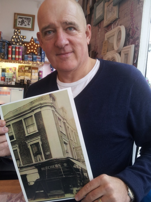  Ian Morris with a 1904 photo of his shop. Back then it was a butcher’s shop. Even now the pink-fronted Map Gift Shop on Junction Road has butchers’ hooks.